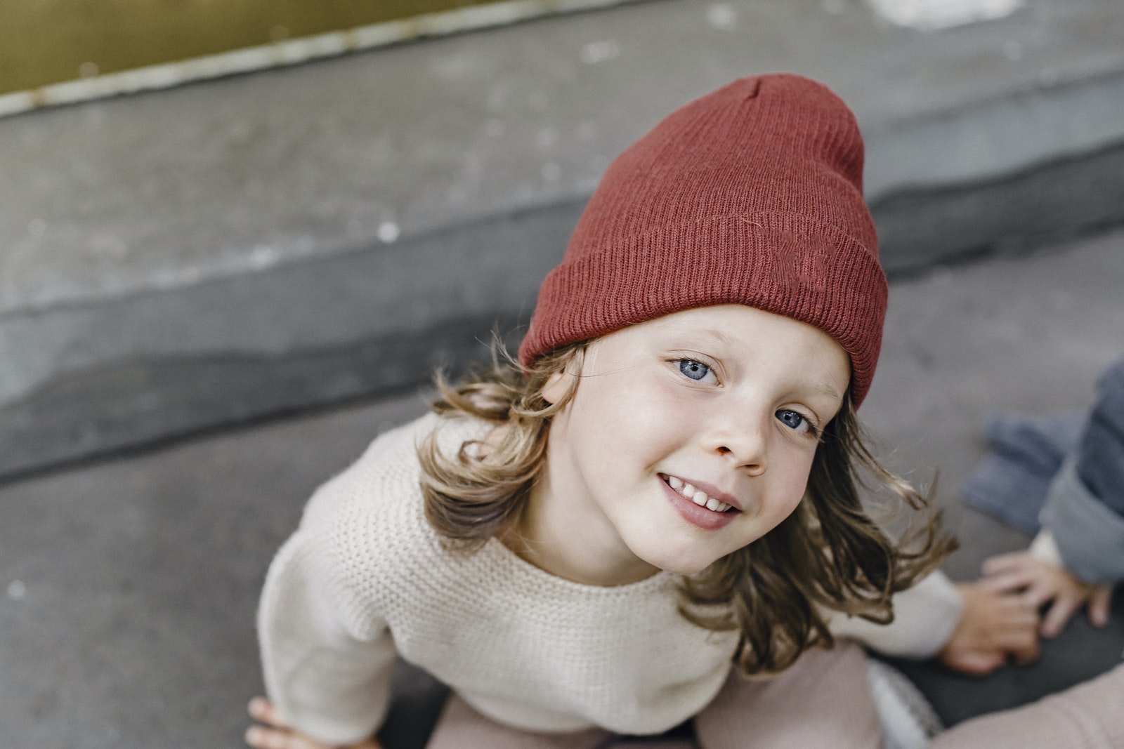 From above of cheerful girl in hat looking at camera while sitting on doorstep with anonymous kid on blurred background