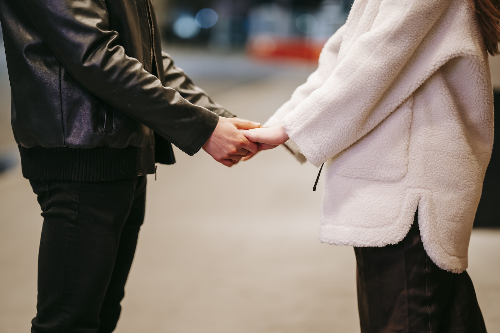 Side view of unrecognizable couple in warm clothes holding hands while standing on blurred background during romantic date on evening time